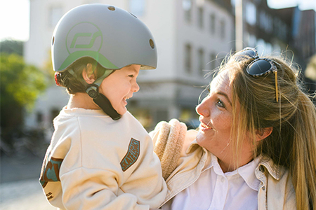 Mutter hat Kind auf dem Arm, beide lachen, das Kind trägt einen blauen Helm