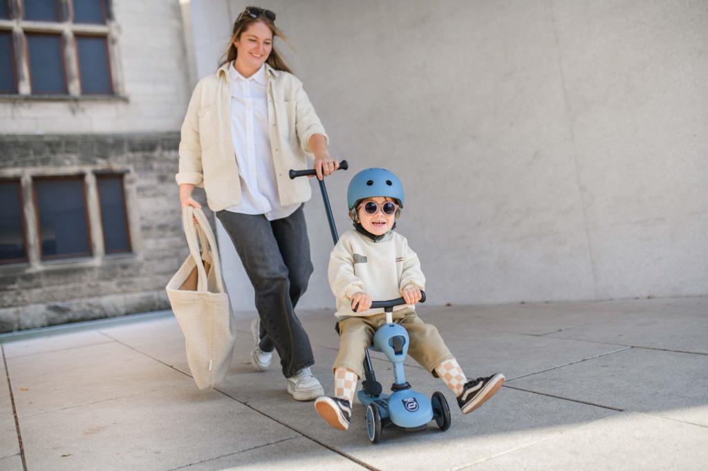 Eine erwachsene Frau schiebt ihren Sohn auf einem Roller mit Sitz, der Roller hat eine Schiebestange, der Junge trägt einen blauen Helm und eine Sonnenbrille