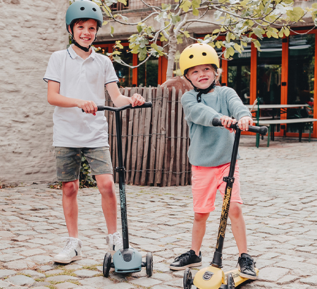 Zwei Juhgs stehen neben ihren Scootern in einem Hinterhof, die Scooter sind blau und gelb, die Kinder tragen Helme in der dazu passenden Farbe