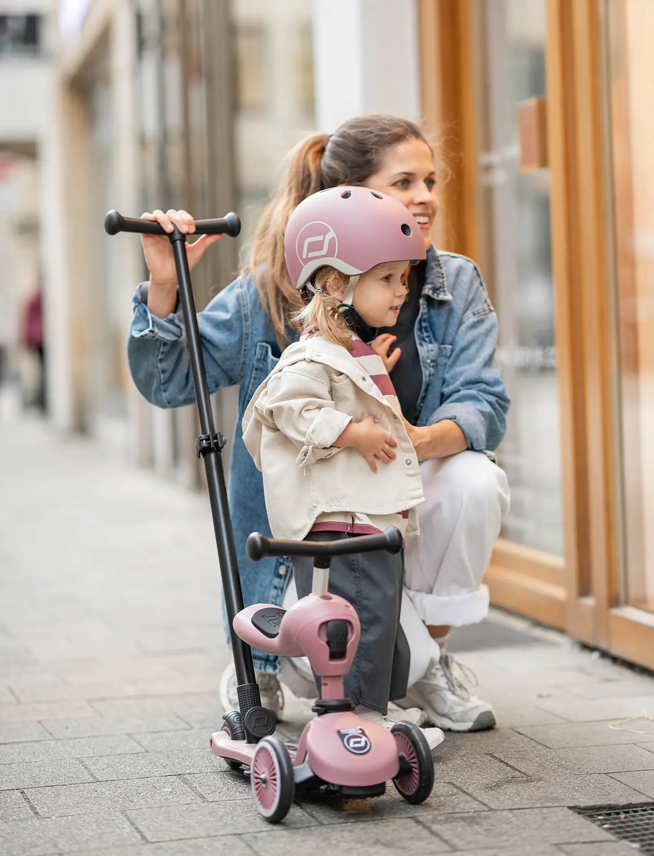 Mutter steht mit Tochter vor Schaufenster, neben den beiden steht ein Highwaykick 1 Push and Go Kinderroller