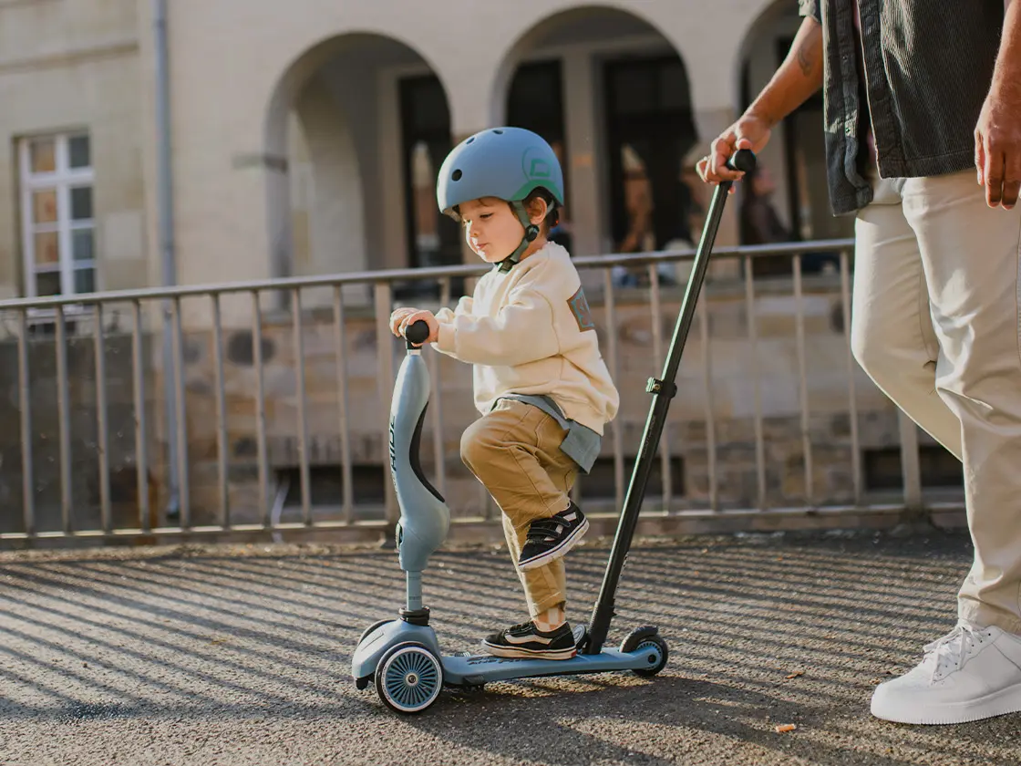 Junge steht auf einem blauen Kinderroller der von seinen Eltern mit Schiebestange geschoben wird