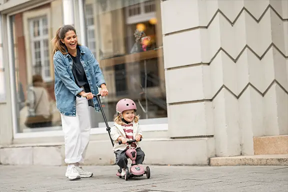 A brown-haired mother pushes her little blonde daughter on a Push and Go scooter in the color Wildberry