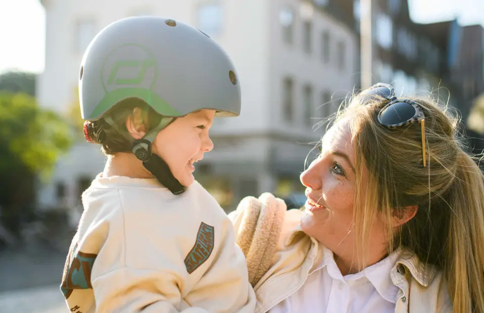Mutter hält kleinen Jungen am Arm, beide lachen sich an, der Junge trägt einnen blauen Helm