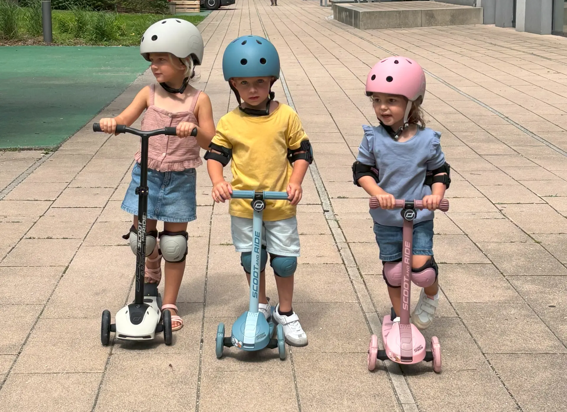 Two girls and a boy are standing next to each other on their scooters, wearing helmets.